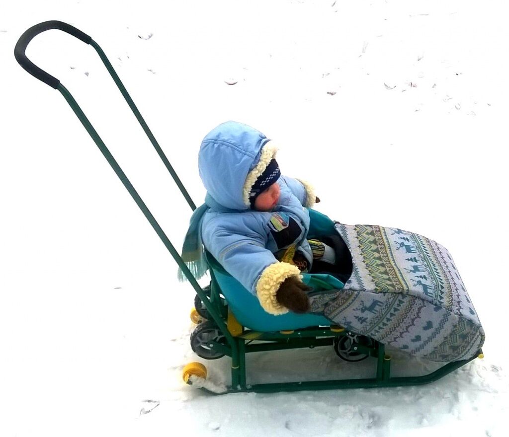 на фото Санки Тимка 5 универсал с сидящим в зимнем комбинезоне ребенком с накидкой попонкой, укрывающей ножки